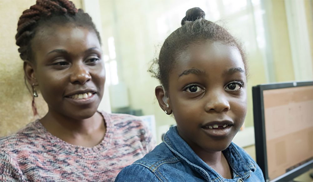 Tobile sits on her mother’s lap, with a hearing aid visible in her ear. They are both smiling.