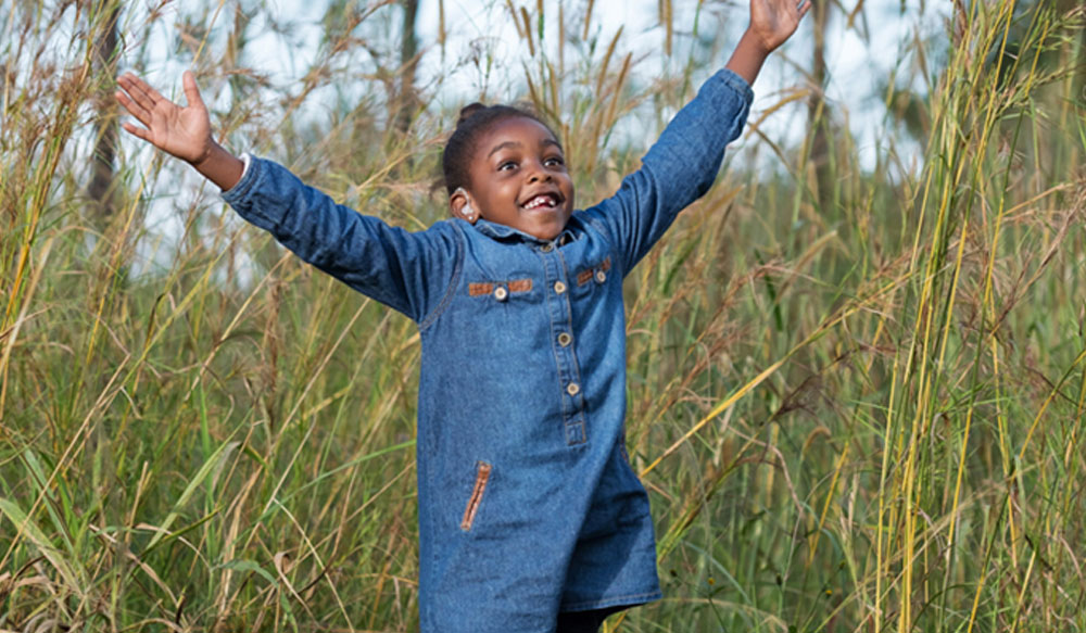 Tobile is outdoors surrounded by long grass, her arms thrown in the air, a radiant smile on her face